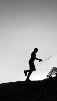 Silhouette of a man exercising with a jump rope on a hill during sunset, creating a serene outdoor scene.